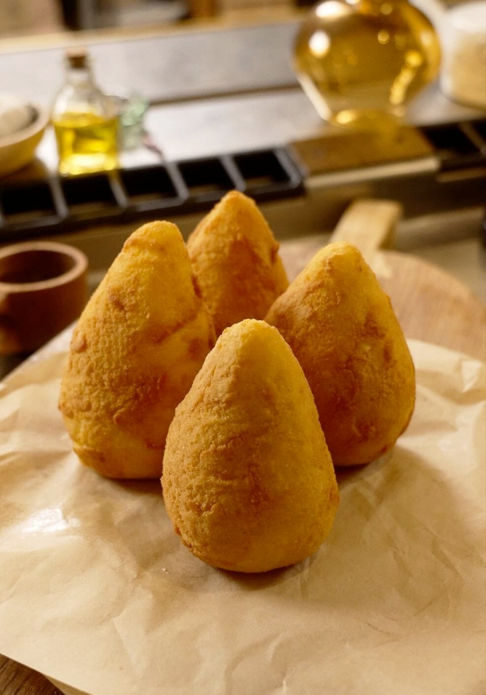 Four golden, cone-shaped fried rice balls sit on parchment paper, placed on a wooden board in a kitchen setting.