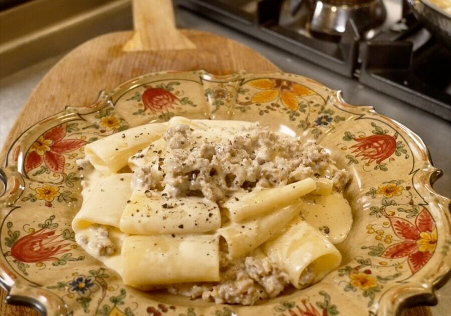 A decorative plate of pasta topped with a creamy meat sauce sits on a wooden cutting board near a gas stove in a kitchen.