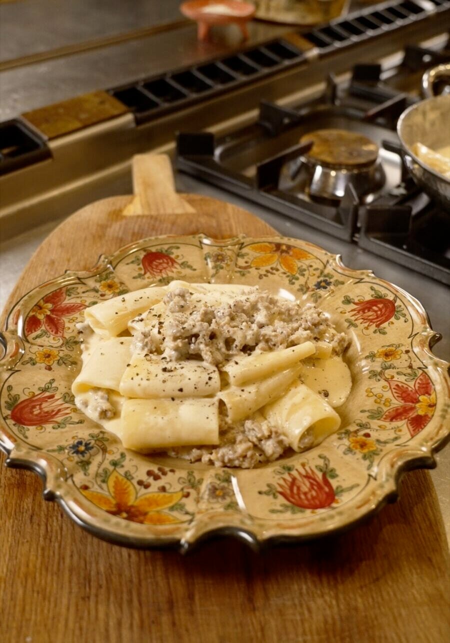 A decorative plate of pasta topped with a creamy meat sauce sits on a wooden cutting board near a gas stove in a kitchen.