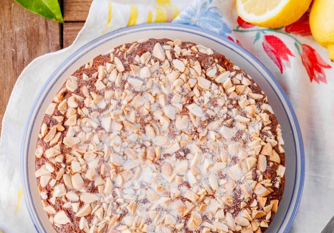 Round cake topped with chopped almonds on a plate, surrounded by lemons on a floral tablecloth.