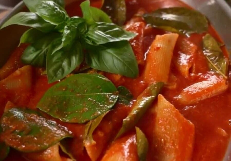 A pot of penne pasta in tomato sauce with basil leaves and vegetables, next to a lemon and a teacup on a wooden table.