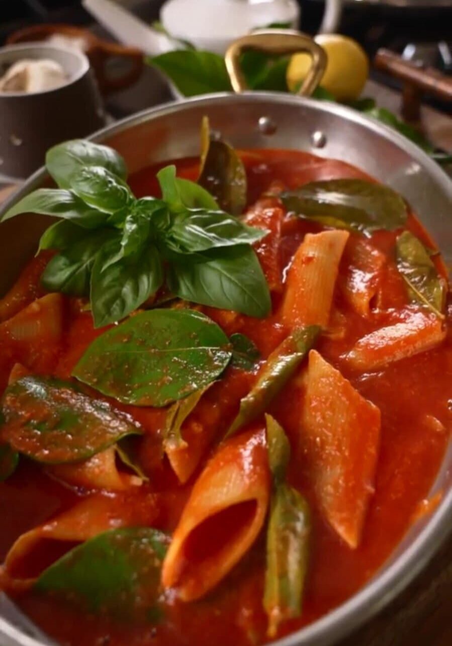 A pot of penne pasta in tomato sauce with basil leaves and vegetables, next to a lemon and a teacup on a wooden table.