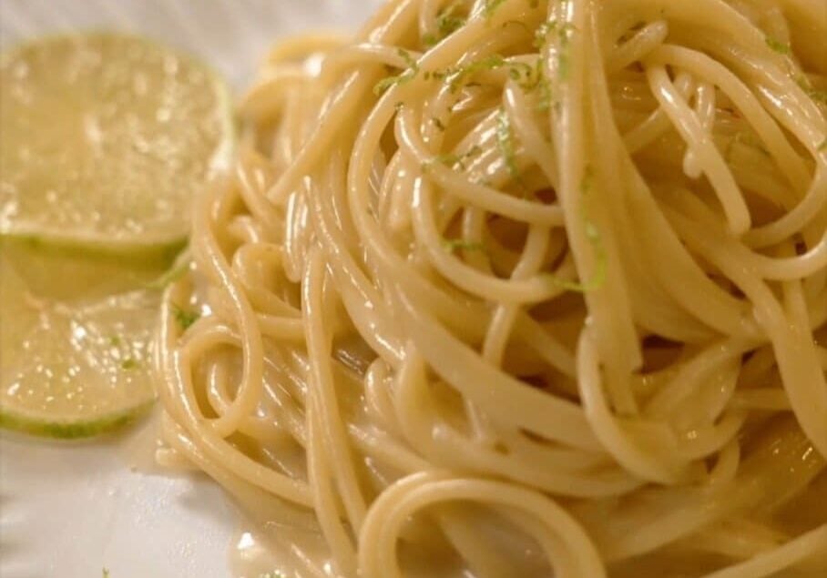 A plate of spaghetti noodles topped with grated lime zest, accompanied by two lime slices on the side.