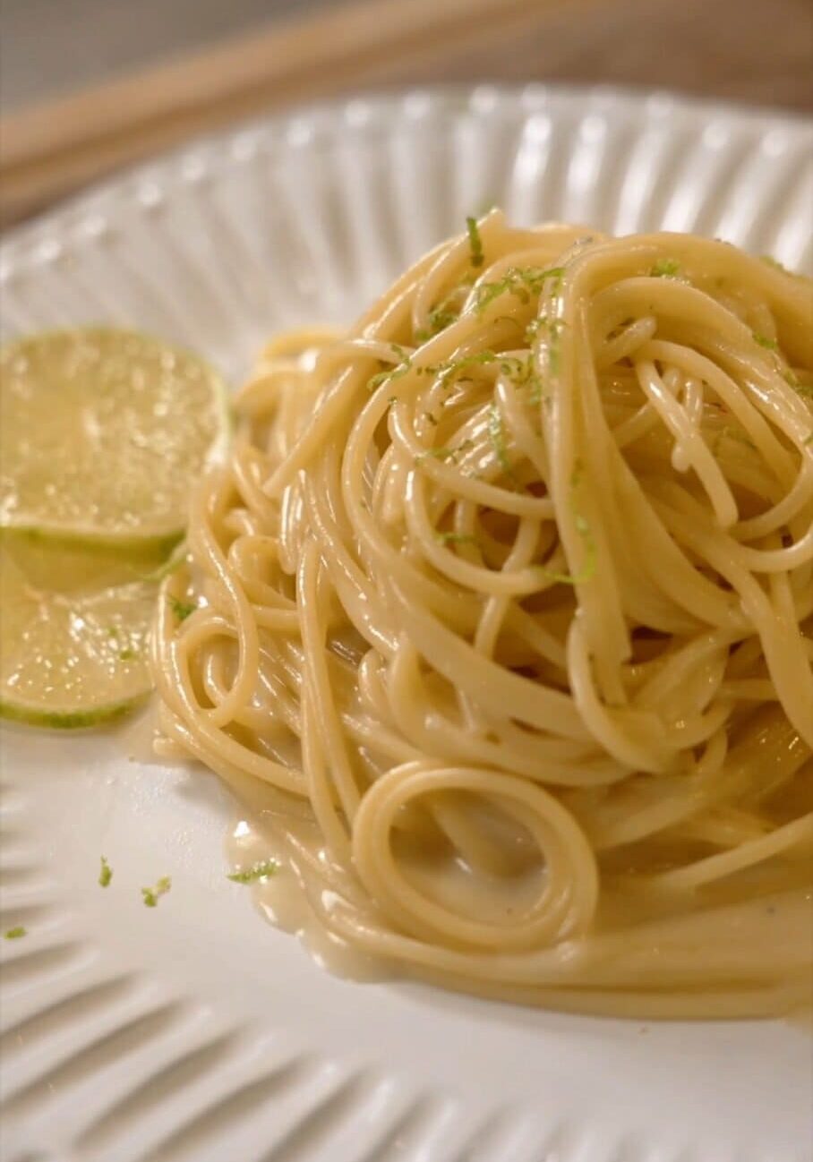 A plate of spaghetti noodles topped with grated lime zest, accompanied by two lime slices on the side.