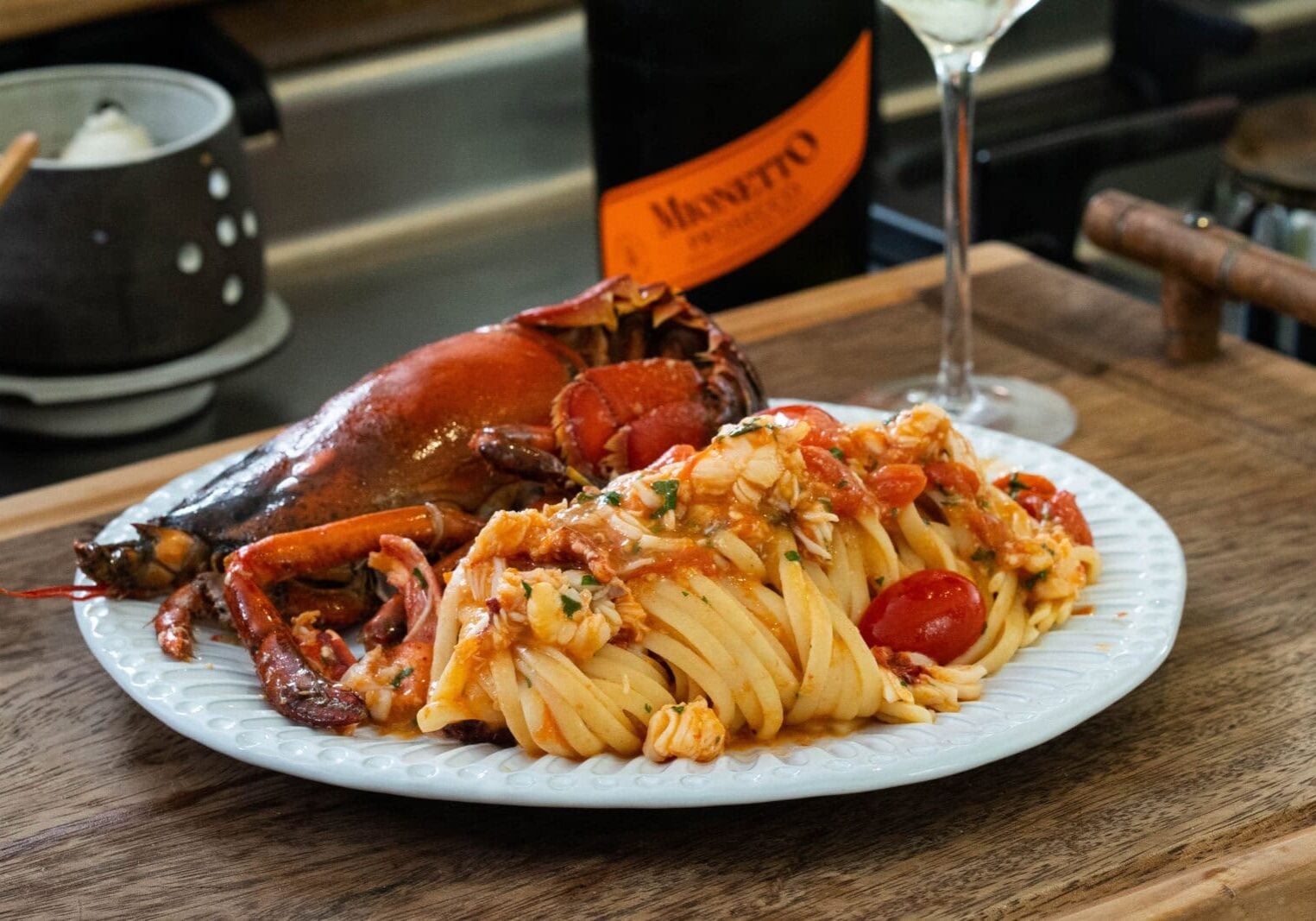 A plate of pasta with cherry tomatoes and lobster in a tomato-based sauce, served with a bottle of Mionetto and a wine glass on a wooden surface.