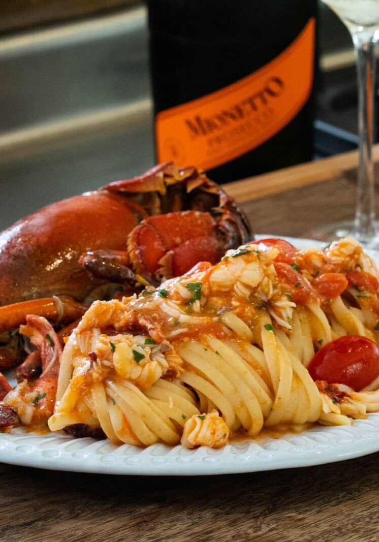 A plate of pasta with cherry tomatoes and lobster in a tomato-based sauce, served with a bottle of Mionetto and a wine glass on a wooden surface.