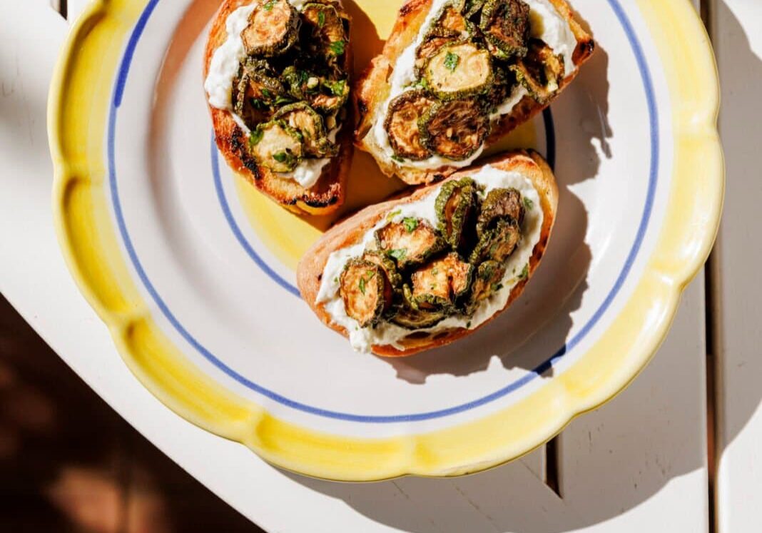 Three slices of bread topped with ricotta and roasted Brussels sprouts on a yellow-rimmed plate, placed on a white table with sunlight casting shadows.