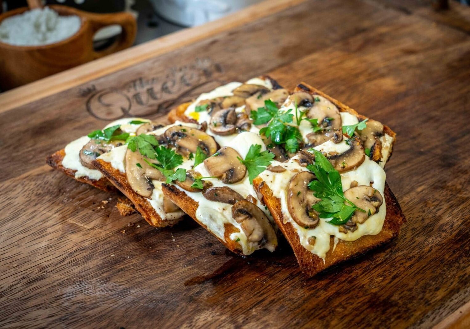Four slices of toasted bread topped with melted cheese, sautéed mushrooms, and parsley on a wooden board.
