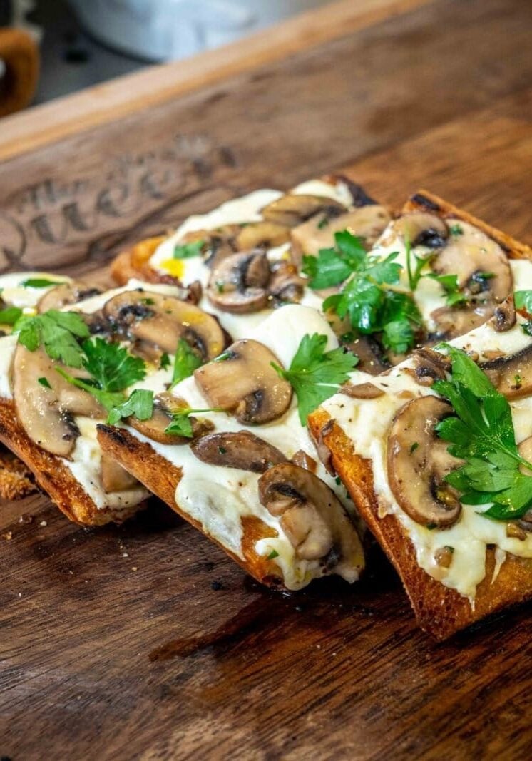 Four slices of toasted bread topped with melted cheese, sautéed mushrooms, and parsley on a wooden board.