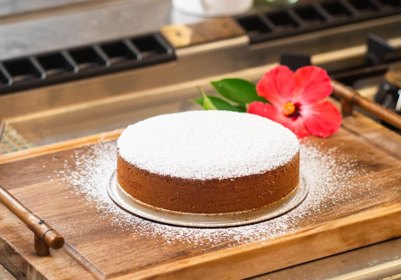 A round cake dusted with powdered sugar sits on a wooden board in a kitchen. A red hibiscus flower and green leaves are placed next to it.