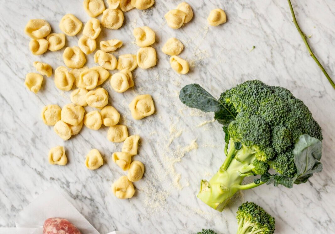 Pasta, broccoli, sausages, garlic, and red chili peppers are arranged on a marble surface.