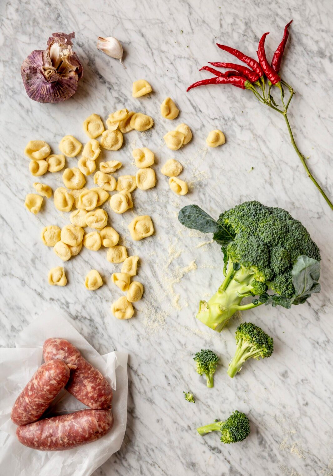 Pasta, broccoli, sausages, garlic, and red chili peppers are arranged on a marble surface.