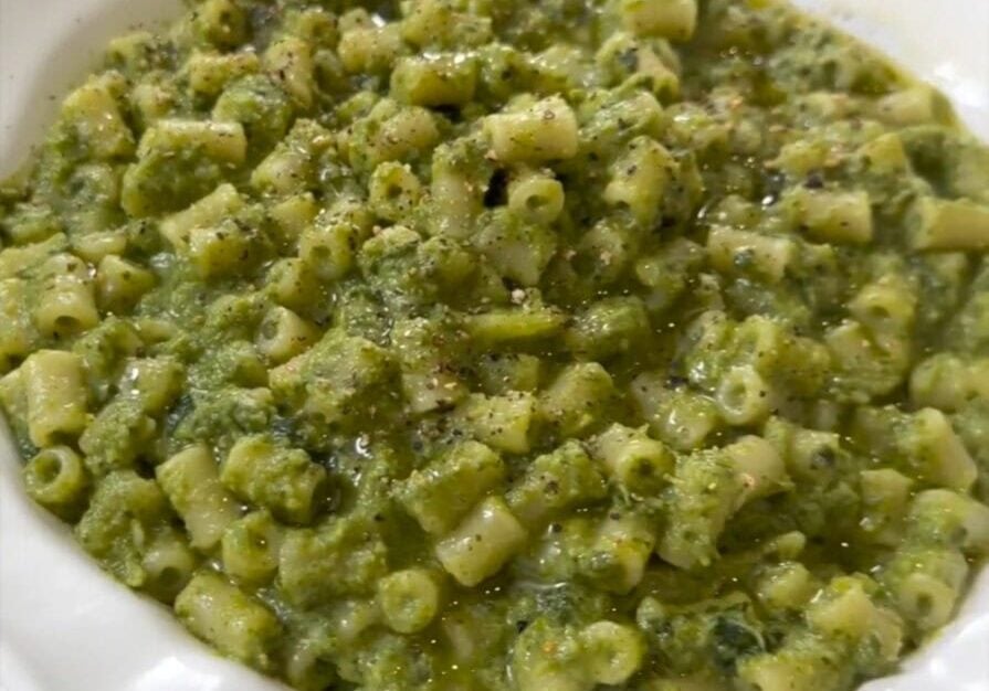 A white bowl filled with pasta mixed in a green pesto sauce, garnished with black pepper and olive oil, placed on a wooden table.
