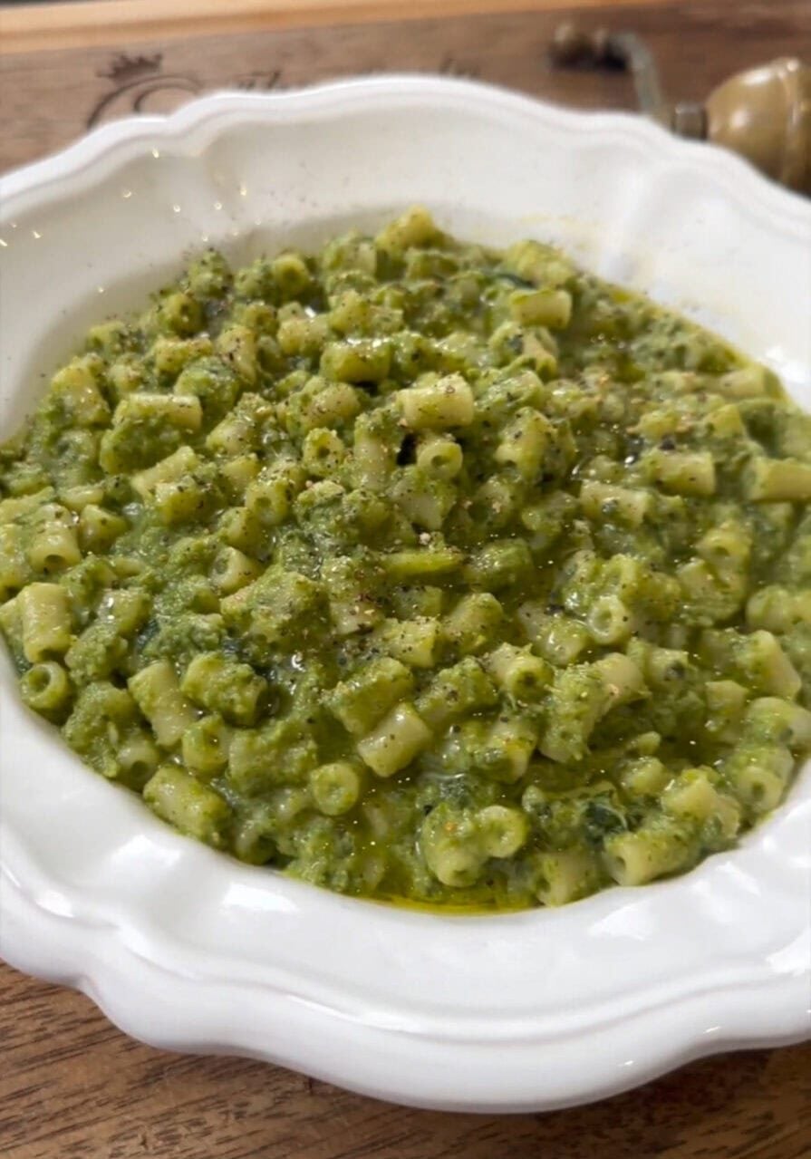 A white bowl filled with pasta mixed in a green pesto sauce, garnished with black pepper and olive oil, placed on a wooden table.