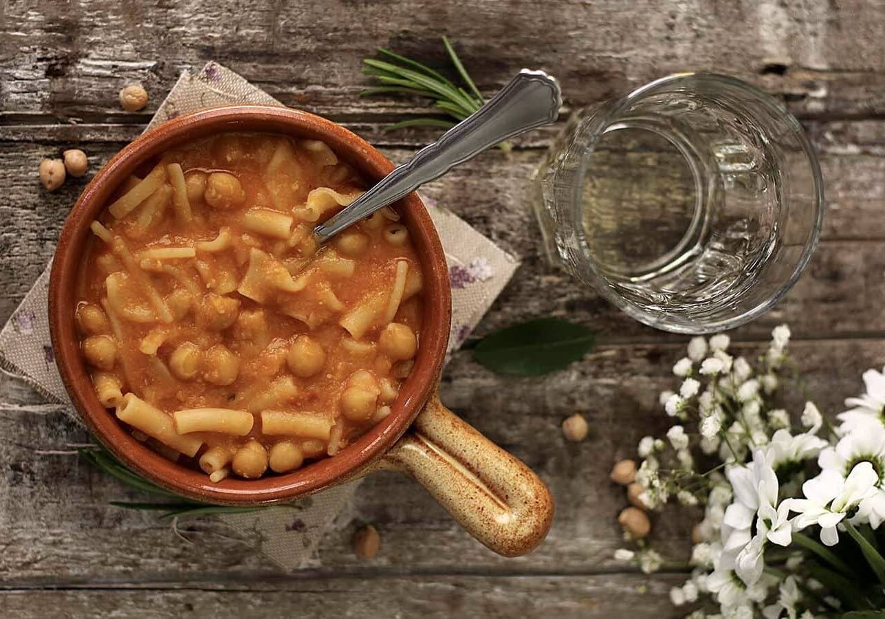 A bowl of pasta with chickpeas and tomato sauce on a rustic wooden surface. A spoon rests in the bowl. Nearby are a glass of water, white flowers, rosemary, and chickpeas.