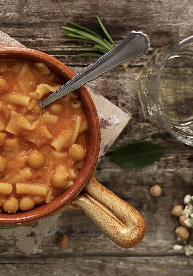 A bowl of pasta with chickpeas and tomato sauce on a rustic wooden surface. A spoon rests in the bowl. Nearby are a glass of water, white flowers, rosemary, and chickpeas.