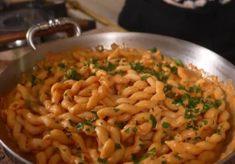 A skillet filled with cooked pasta in a creamy sauce, garnished with chopped parsley. A person in a floral top is visible in the background.