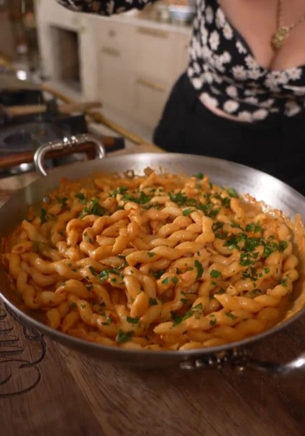A skillet filled with cooked pasta in a creamy sauce, garnished with chopped parsley. A person in a floral top is visible in the background.