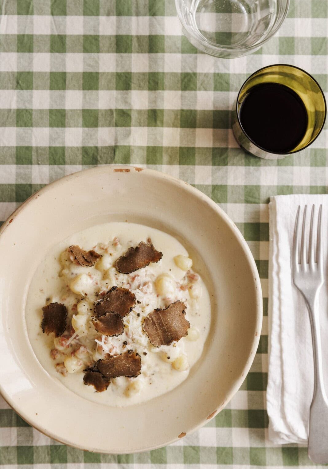 A bowl of creamy gnocchi topped with truffle slices, set on a green and white checkered tablecloth. A glass of water, a glass of red wine, a fork, and a knife are beside it.