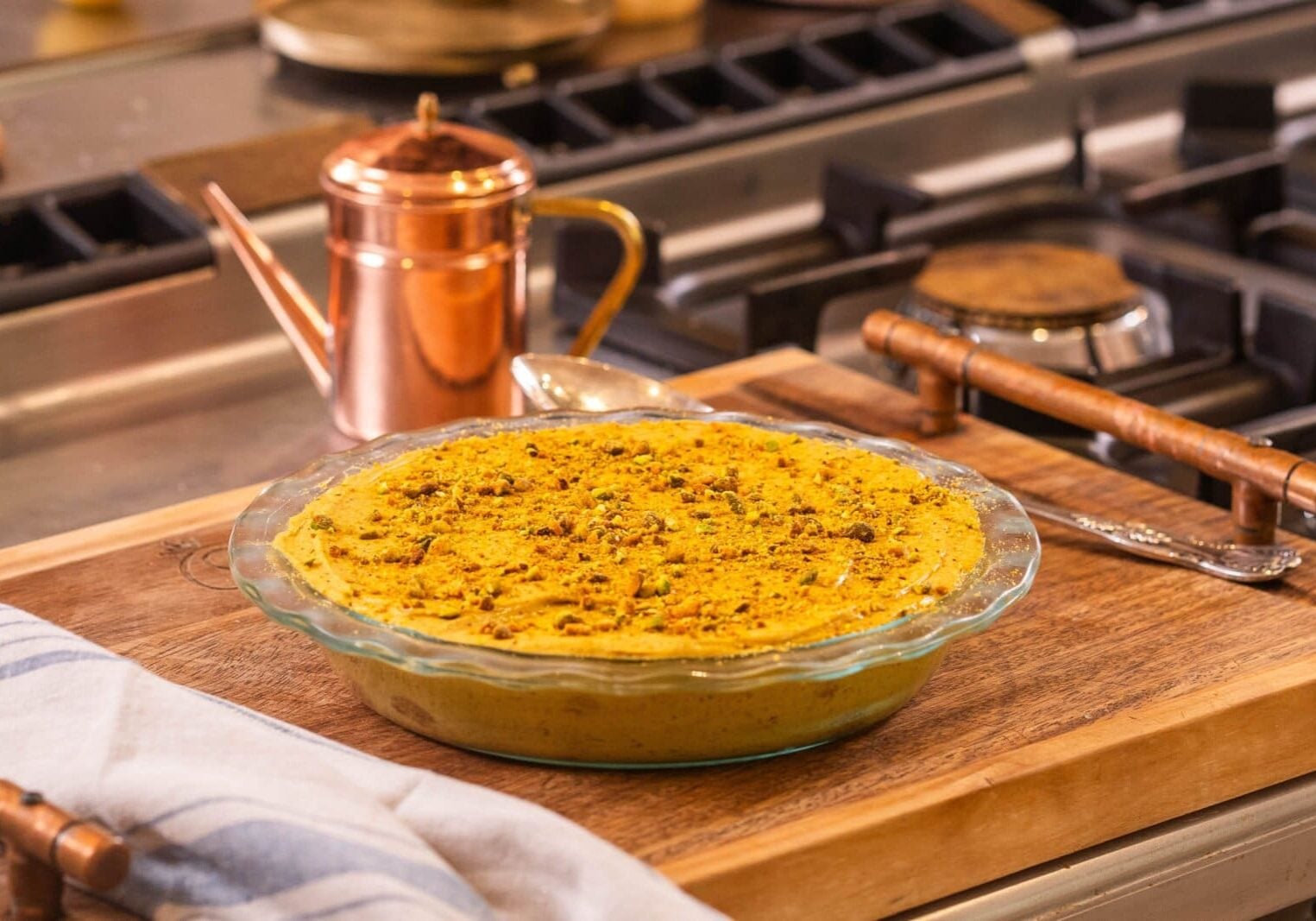 A pie in a glass dish is on a wooden board. It has a yellow filling with a crumbly topping. A copper pot is in the background, and a striped cloth is partially visible in the foreground.