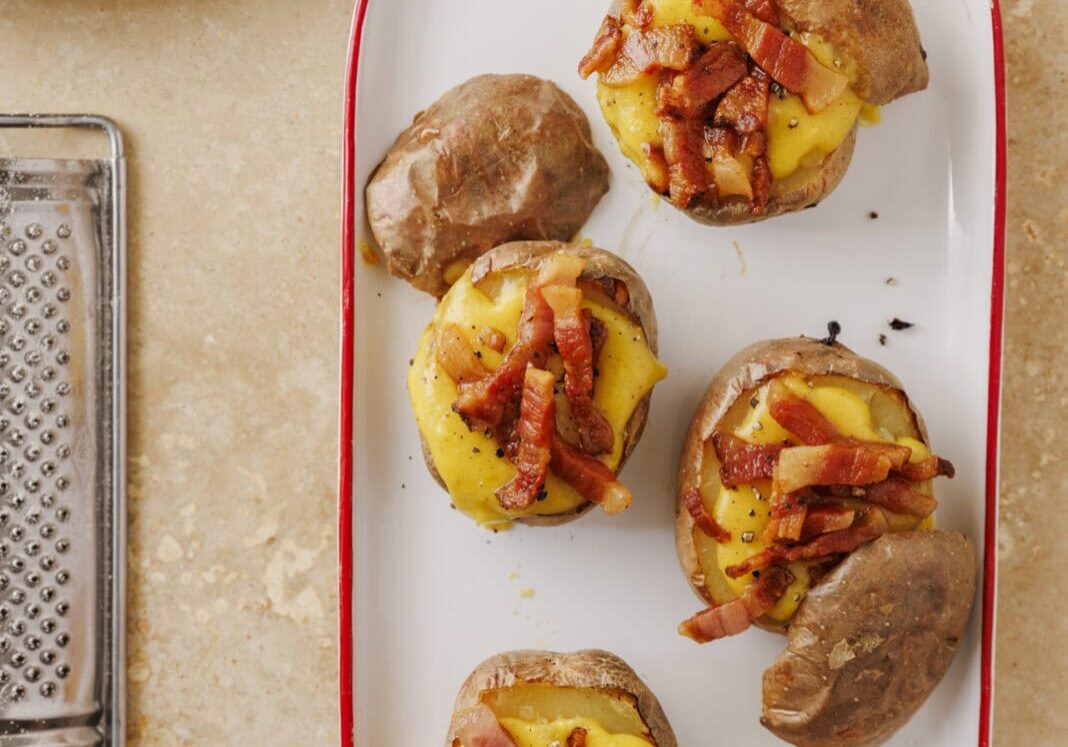 Four baked potatoes topped with melted cheese and bacon pieces on a white rectangular plate. Nearby are eggs, a cheese grater, and a block of cheese on a plate.