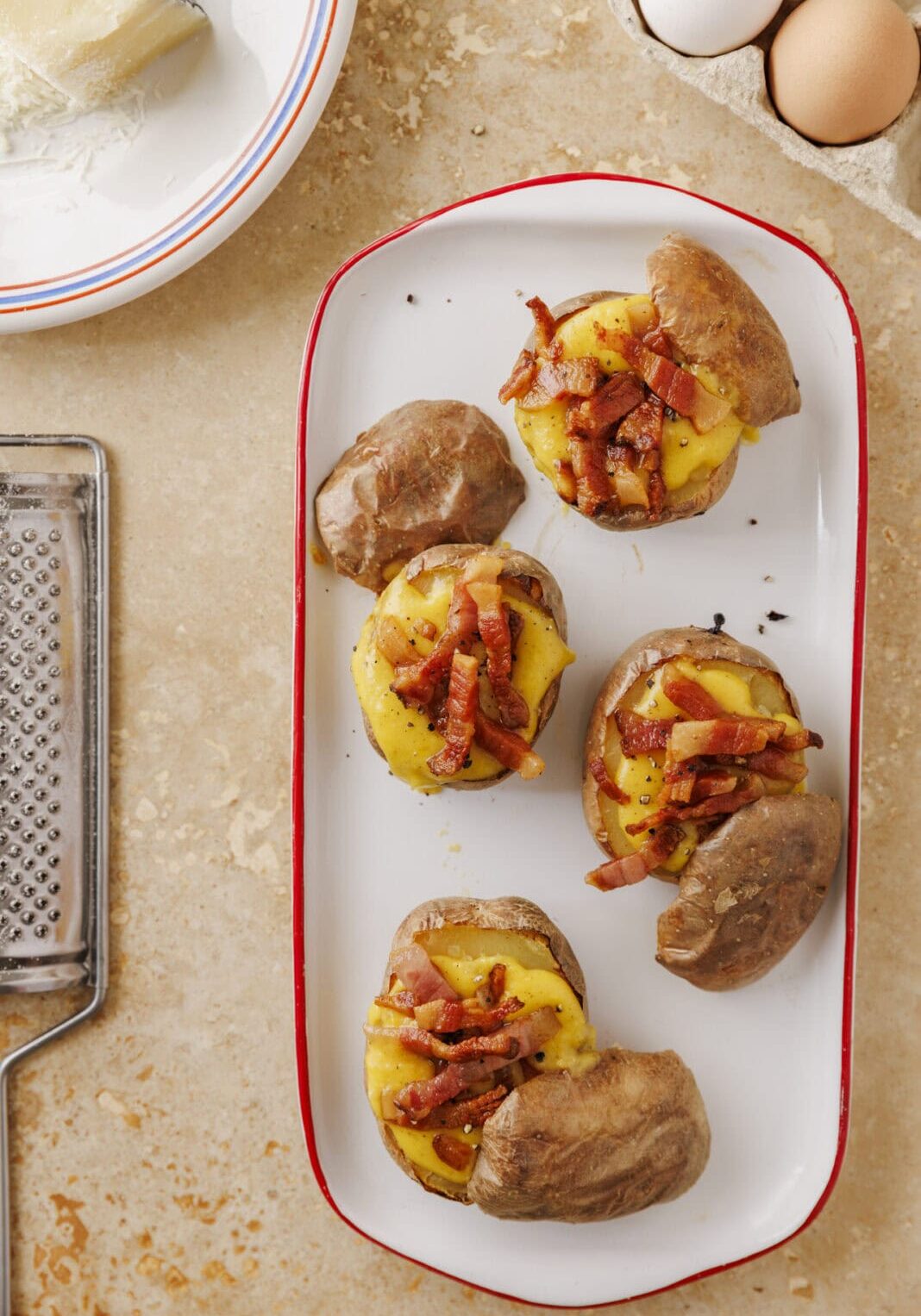 Four baked potatoes topped with melted cheese and bacon pieces on a white rectangular plate. Nearby are eggs, a cheese grater, and a block of cheese on a plate.