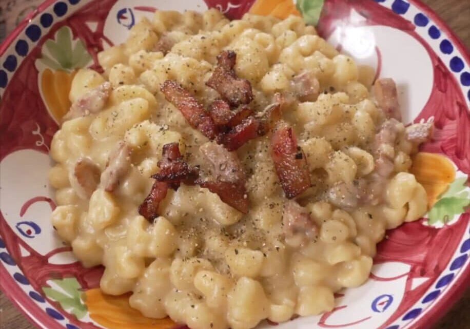 A colorful bowl filled with creamy macaroni and cheese topped with crispy bacon pieces and a sprinkle of black pepper, placed on a wooden surface.