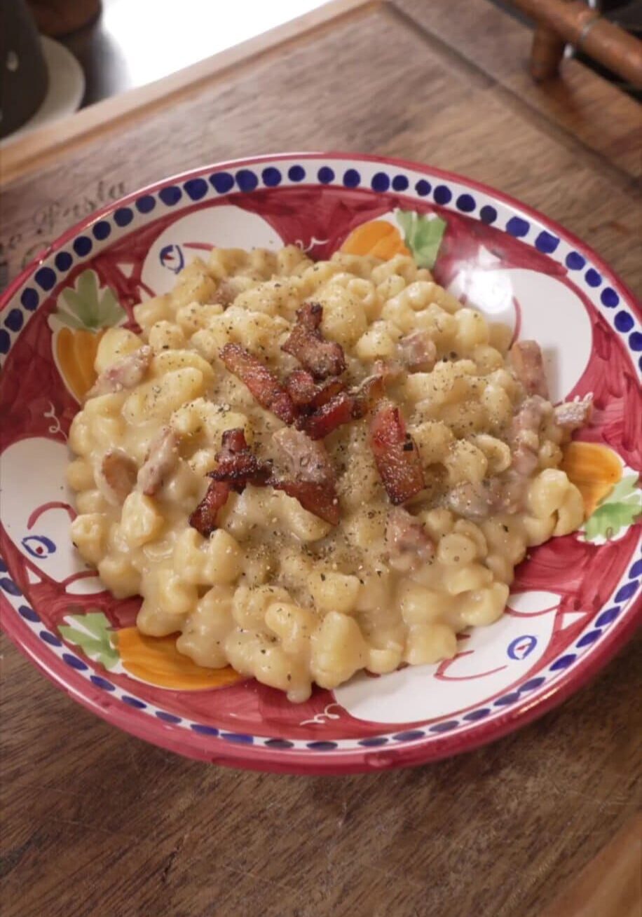 A colorful bowl filled with creamy macaroni and cheese topped with crispy bacon pieces and a sprinkle of black pepper, placed on a wooden surface.