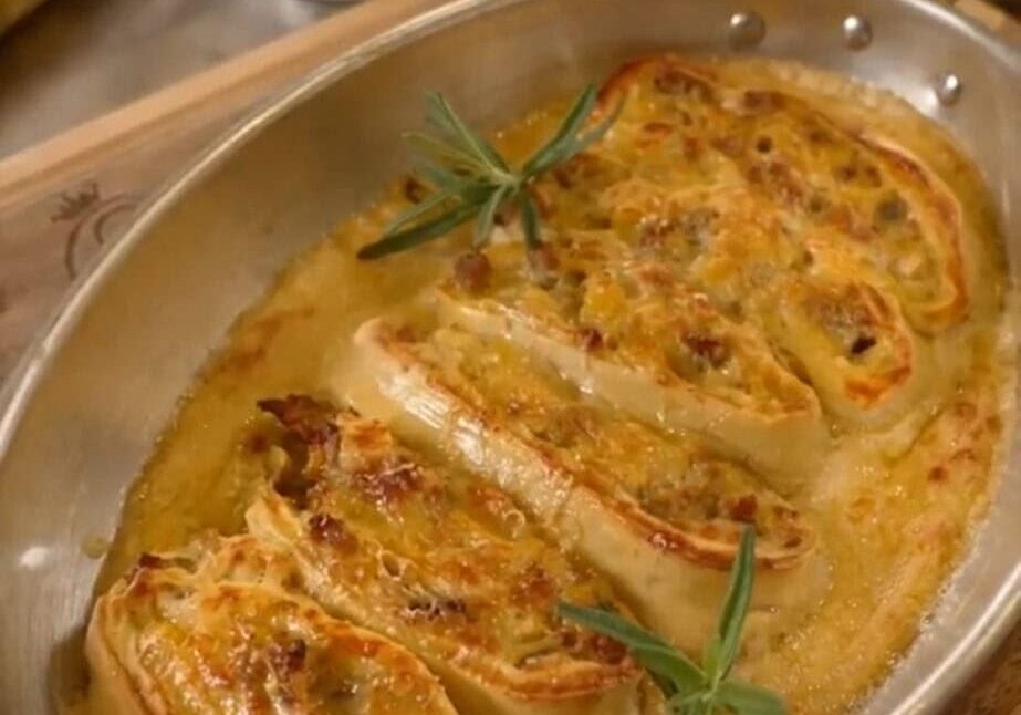 Oval dish with baked stuffed pasta rolls, garnished with rosemary sprigs, on a wooden board. Kettle visible in background.