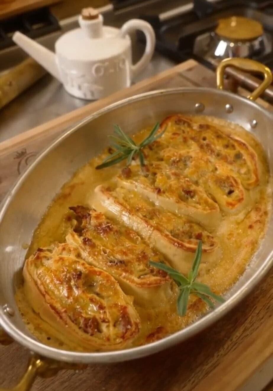 Oval dish with baked stuffed pasta rolls, garnished with rosemary sprigs, on a wooden board. Kettle visible in background.