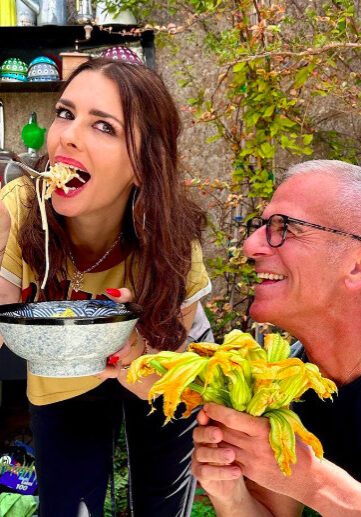 A woman eats pasta from a bowl with a fork while a man next to her holds fresh zucchini flowers and smiles; they are in an outdoor kitchen setting.