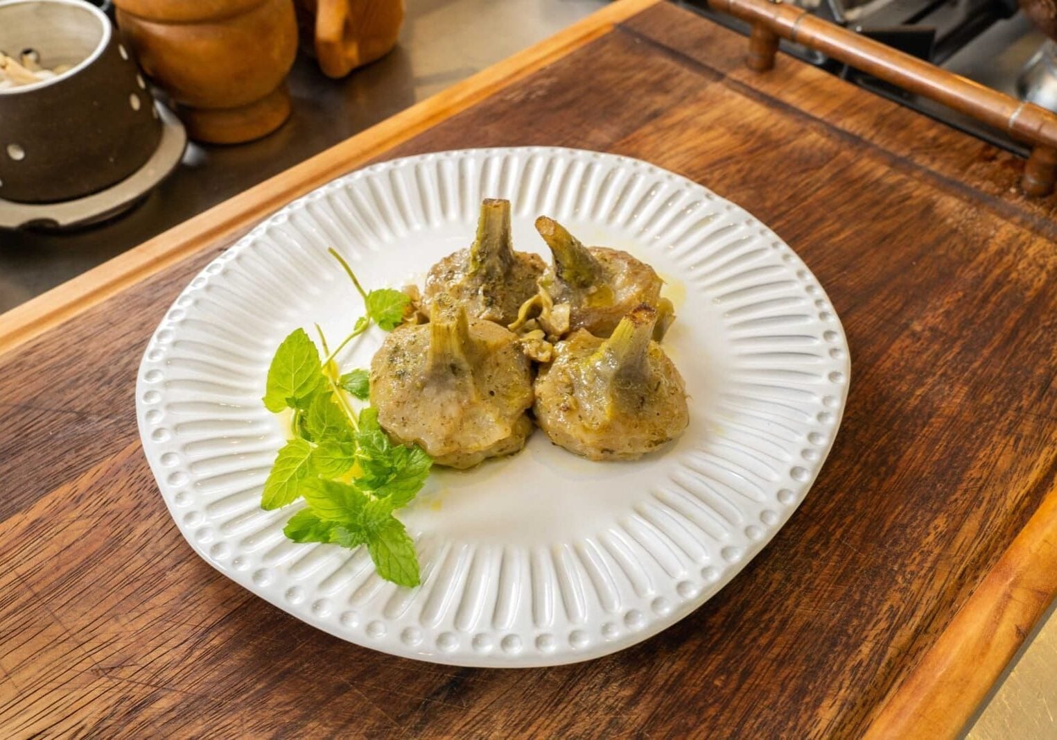 Three cooked artichokes with a garnish of mint leaves on a white plate, placed on a wooden surface.