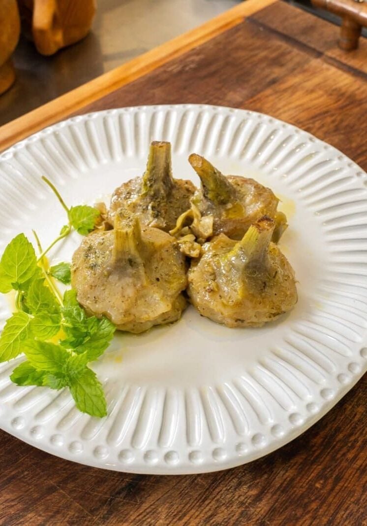 Three cooked artichokes with a garnish of mint leaves on a white plate, placed on a wooden surface.