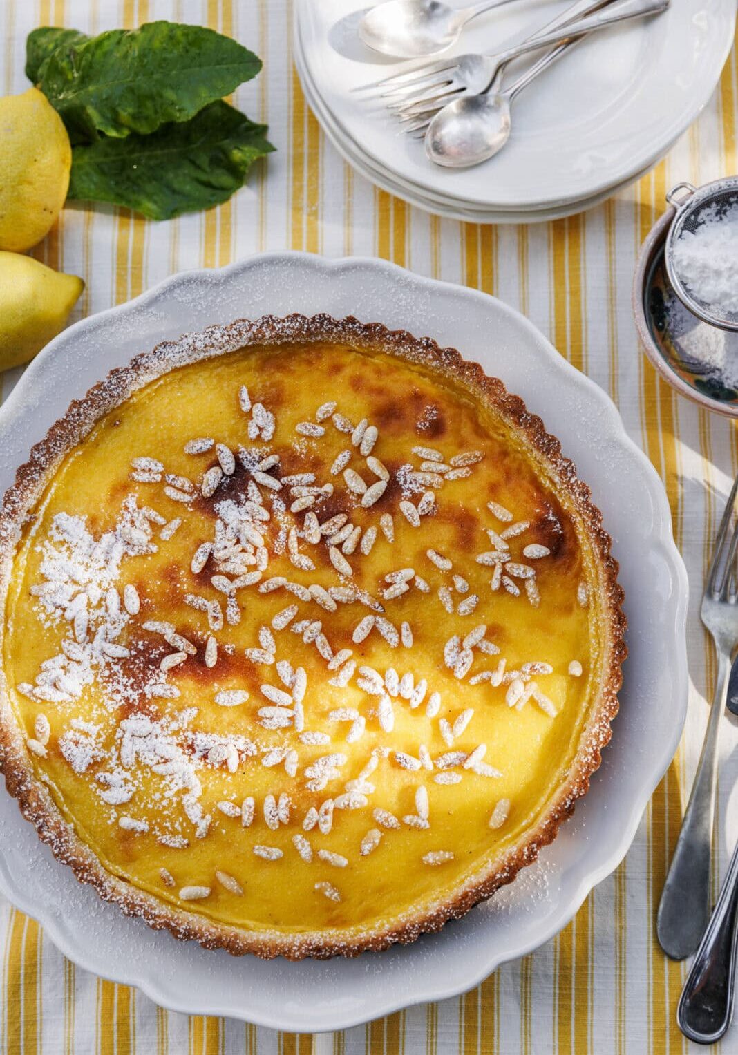 A lemon tart topped with pine nuts and powdered sugar sits on a white plate, surrounded by lemons, mint leaves, cutlery, and stacked plates on a striped tablecloth.