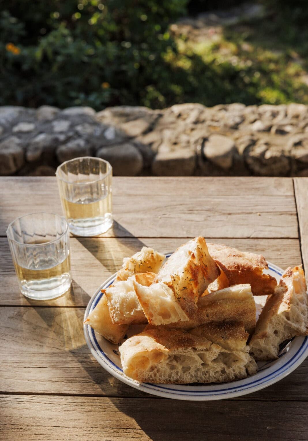 A plate of sliced bread and two glasses of water sit on a wooden table outdoors, with sunlight and greenery in the background.