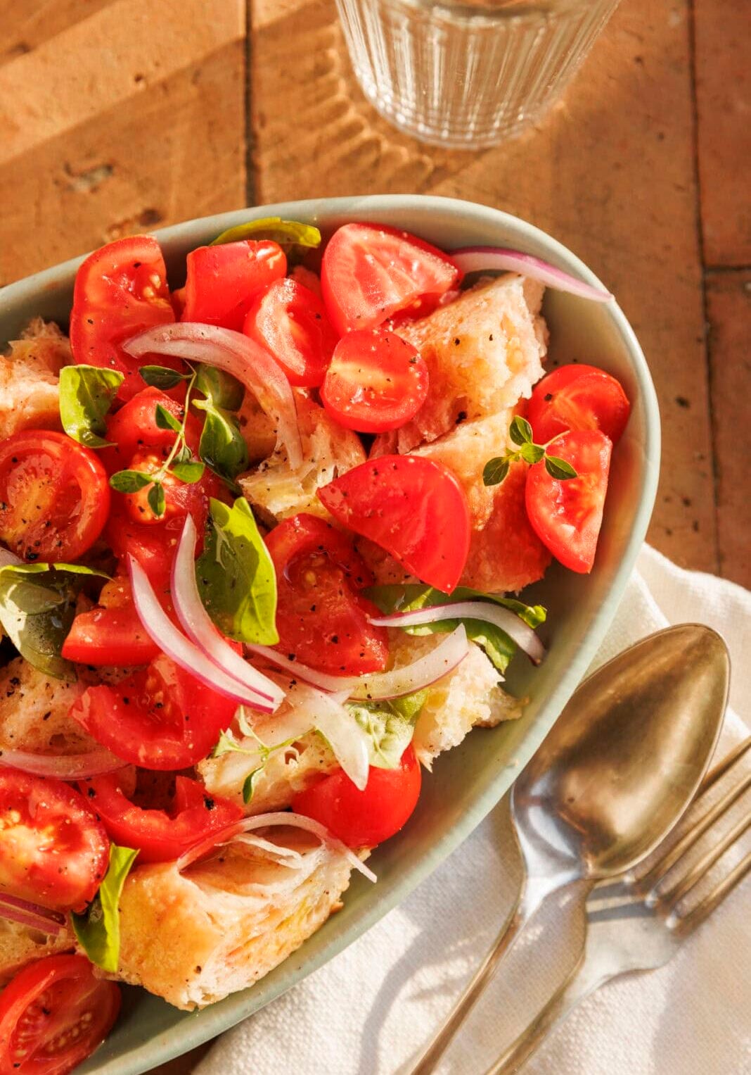 Oval dish with tomato, onion, basil, and bread salad on a napkin beside a spoon, fork, and glass of water on a sunlit table.