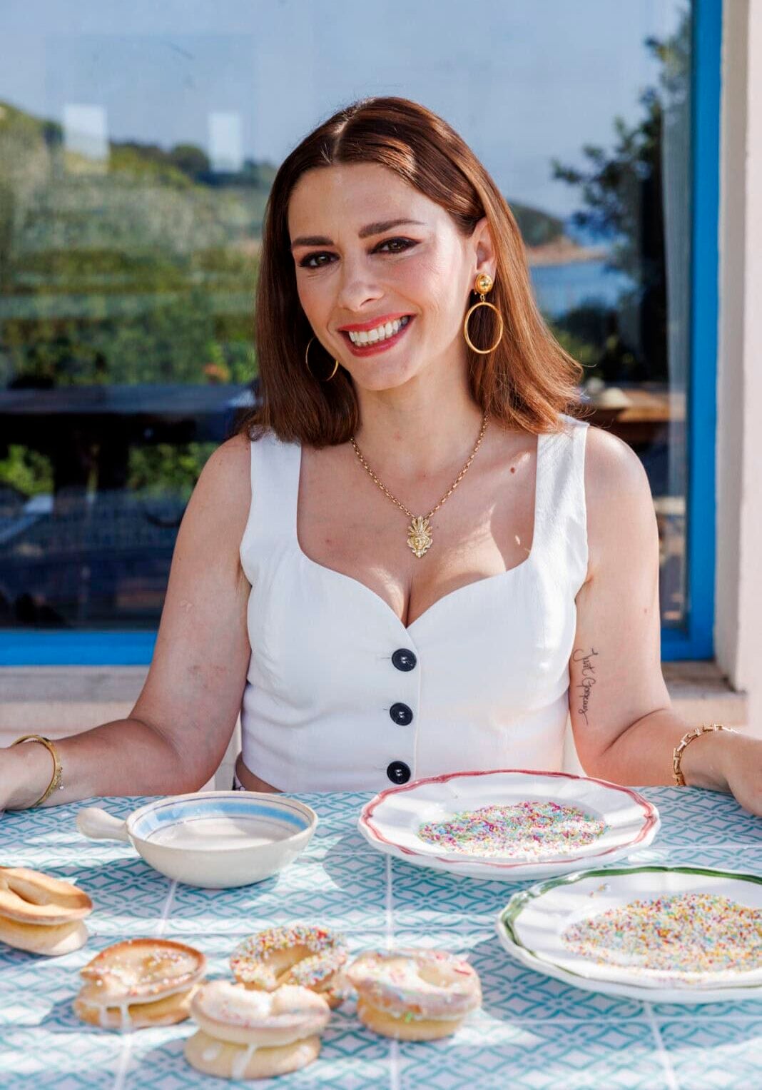 A woman in a white top sits at a table with plates of bagels, bowls of milk, and colorful sprinkles in front of a window on a sunny day.