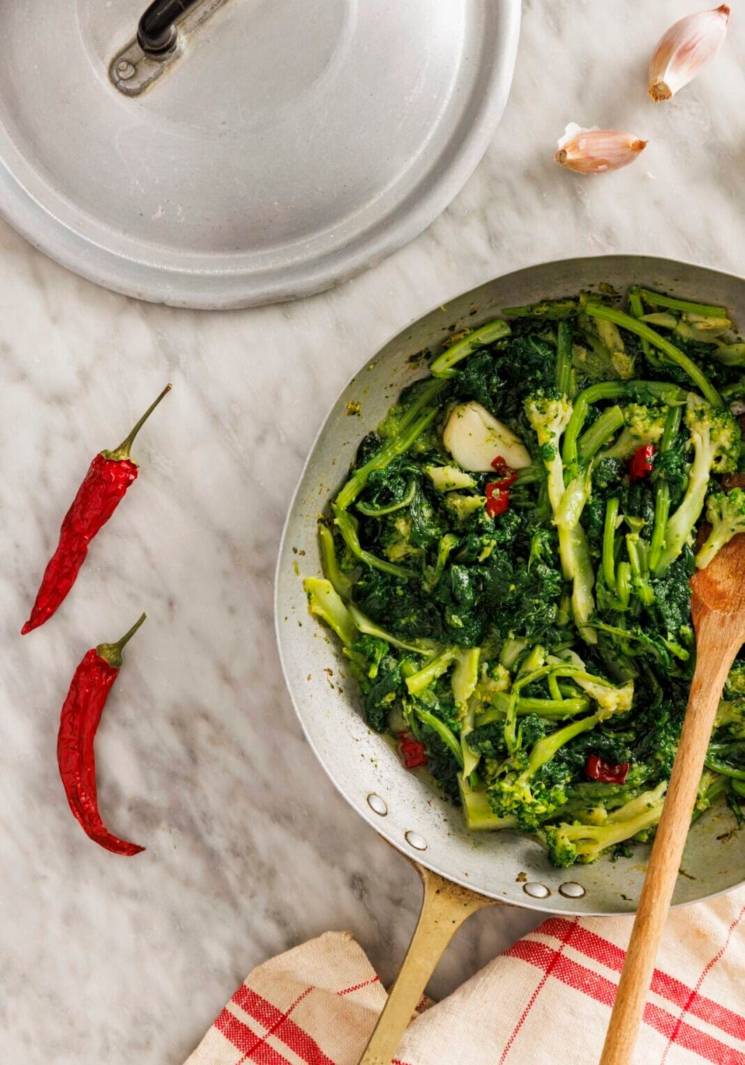 A pan of cooked leafy greens with chopped red chili peppers sits on a marble countertop, surrounded by a pot lid, two garlic cloves, two dried red chilies, and a red-striped towel.