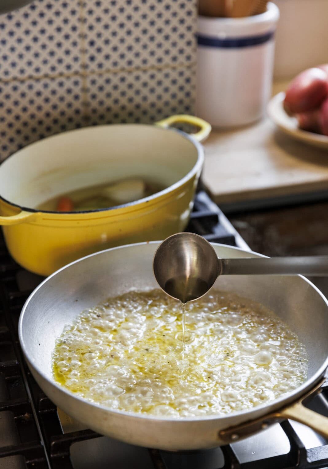 A metal ladle pours liquid into a skillet of bubbling oil on a stovetop, with a yellow Dutch oven and a plate of onions in the background.