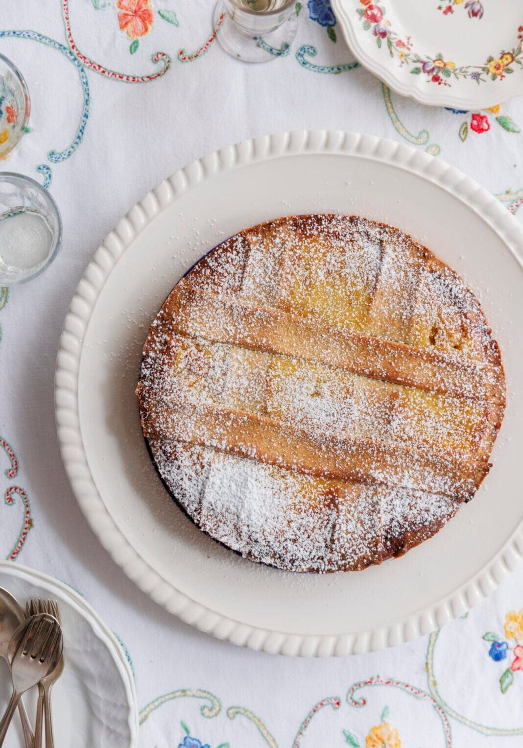 A round cake dusted with powdered sugar on a white platter, set on a table with a white embroidered tablecloth and floral-patterned dishes.