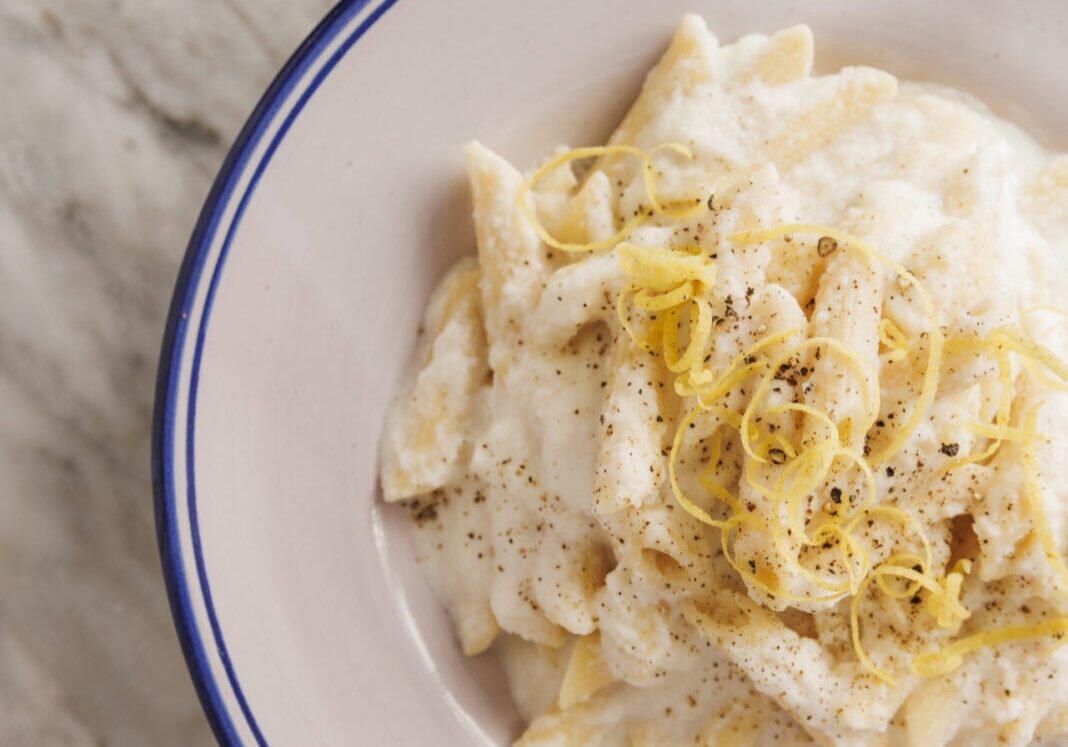 A bowl of pasta in creamy white sauce topped with grated cheese and pepper, garnished with lemon zest, on a marble surface.