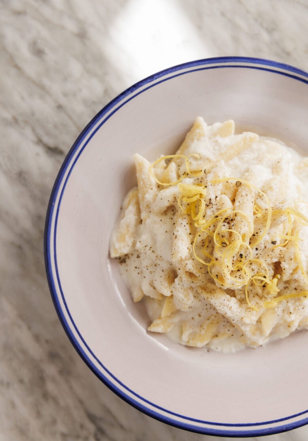 A bowl of pasta in creamy white sauce topped with grated cheese and pepper, garnished with lemon zest, on a marble surface.
