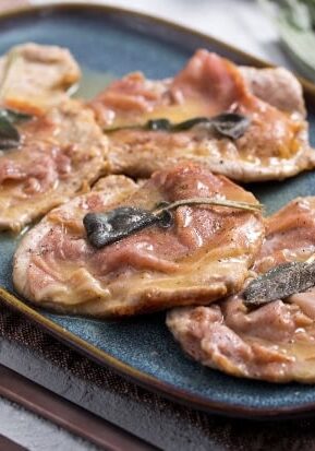 Four pieces of veal saltimbocca topped with prosciutto and sage on a blue oval plate.