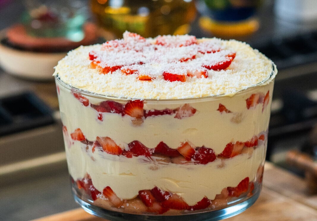 Glass trifle dish with layers of cream, strawberries, and cake, topped with shredded coconut and strawberry slices, on a wooden surface.