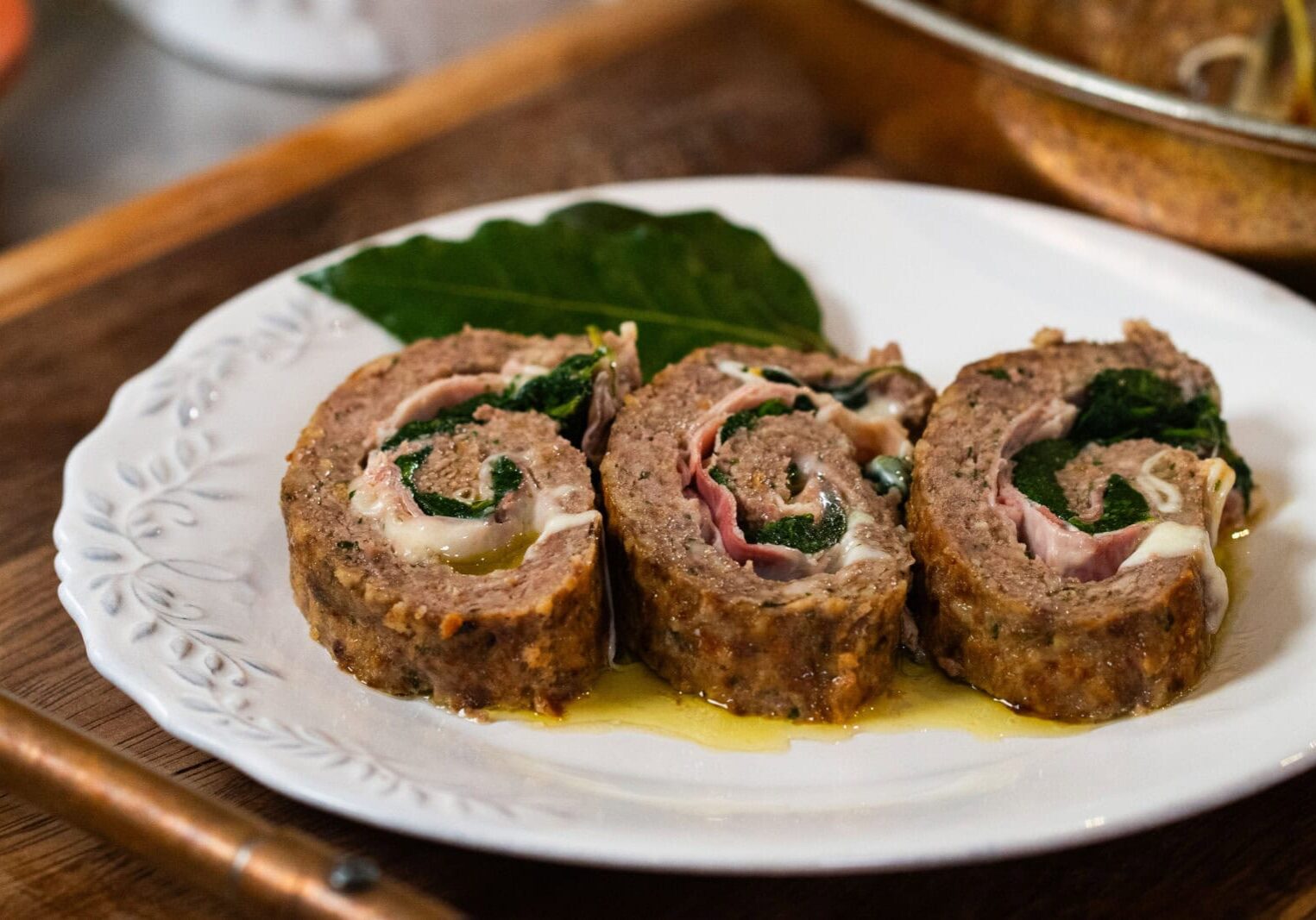 Three slices of meat roulade filled with greens and cheese are arranged on a white plate with a bay leaf garnish.