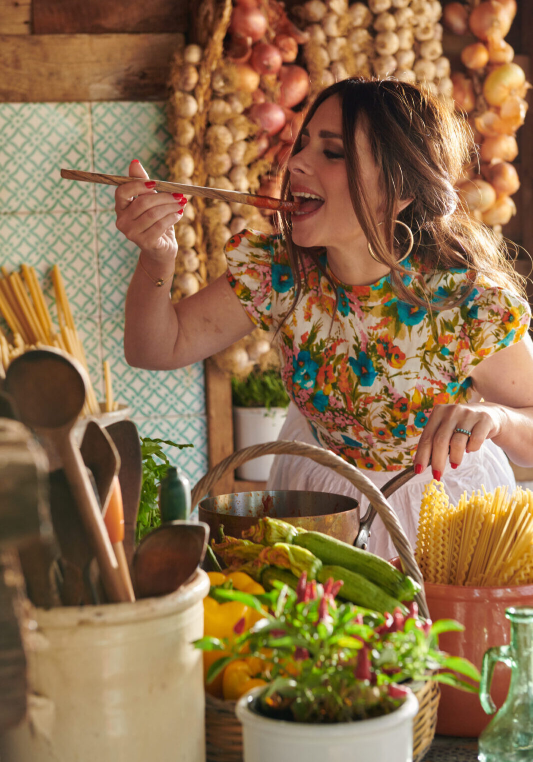 A woman in a floral shirt eats spaghetti with a fork in a rustic kitchen surrounded by fresh vegetables, hanging garlic, and pasta.