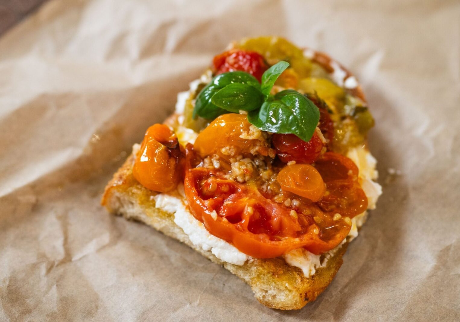 A slice of bread topped with creamy cheese, sliced tomatoes, cherry tomatoes, and fresh basil leaves on parchment paper.