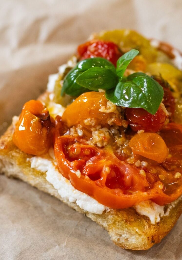 A slice of bread topped with creamy cheese, sliced tomatoes, cherry tomatoes, and fresh basil leaves on parchment paper.