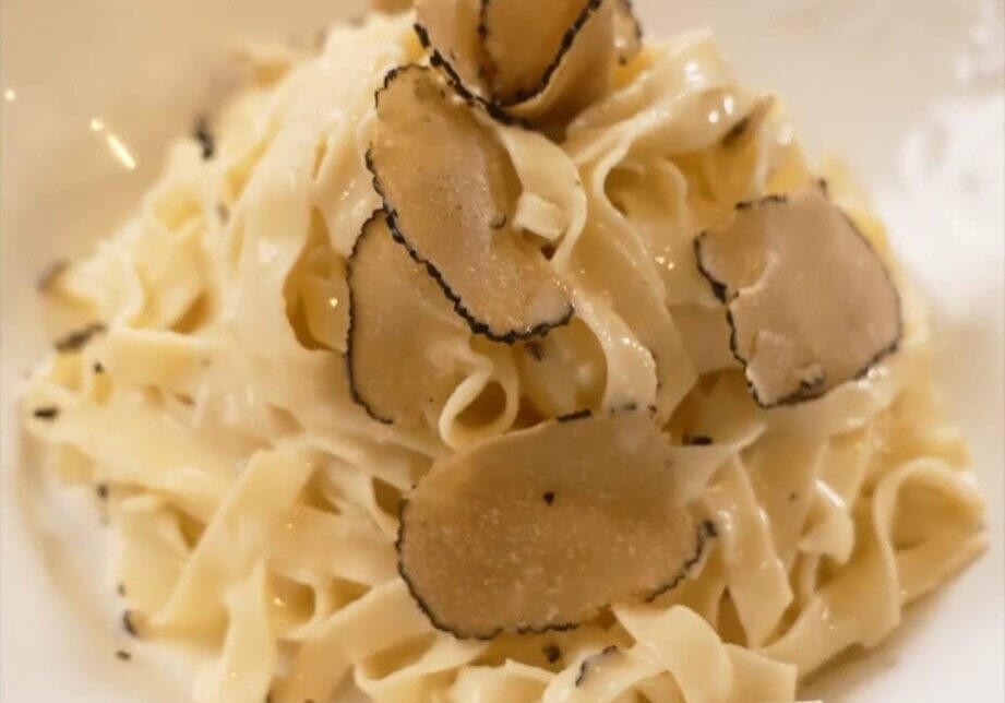 A bowl of fettuccine pasta with cream sauce, topped with thin slices of black truffle on a white plate.