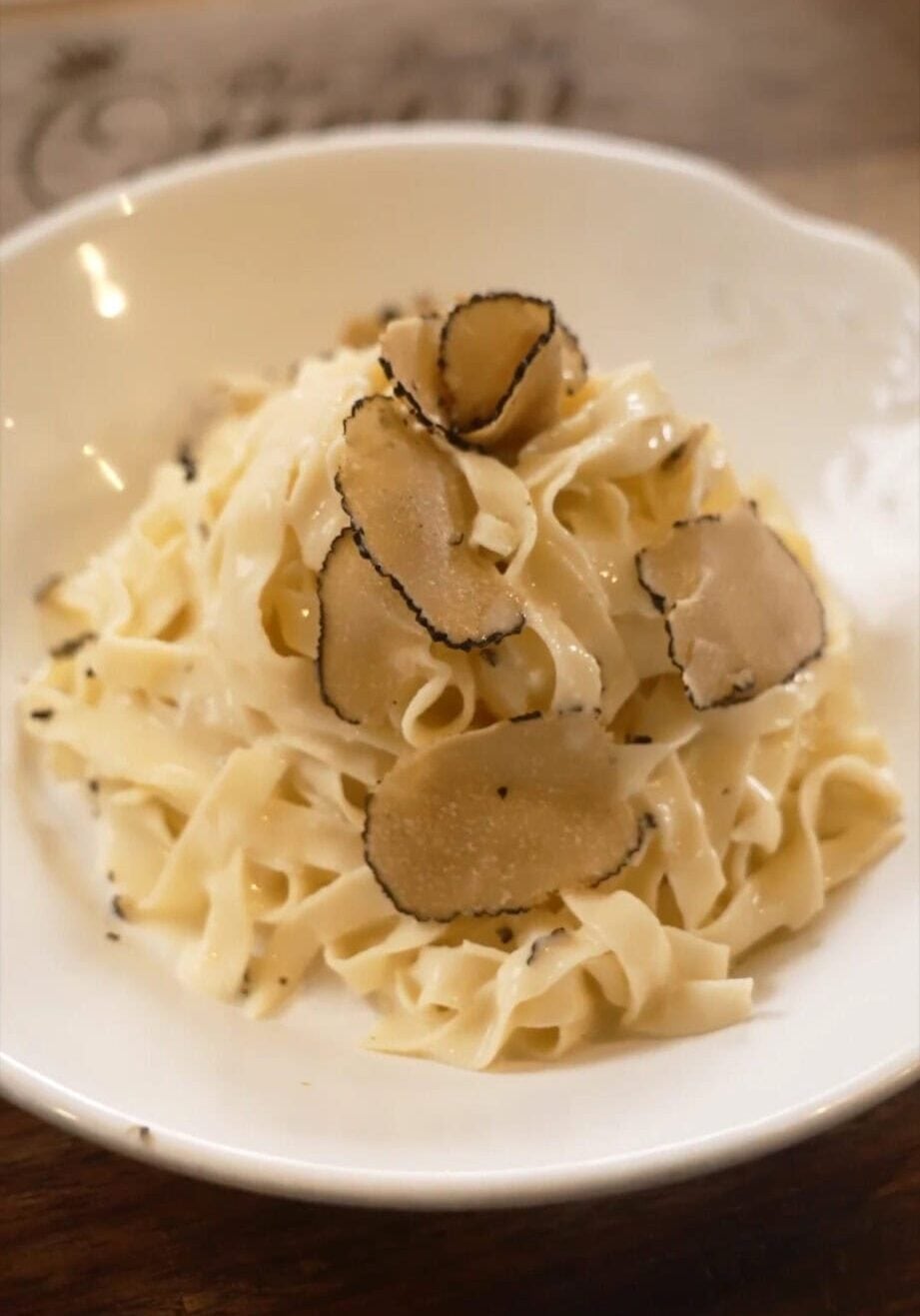 A bowl of fettuccine pasta with cream sauce, topped with thin slices of black truffle on a white plate.